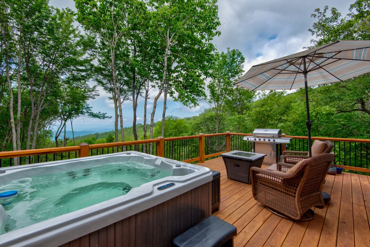 A serene outdoor deck area featuring a bubbling hot tub, grill, outdoor seating, and an umbrella, all oriented to take in the surrounding trees and likely distant views. The scene is peaceful and inviting, suggesting relaxation after a day of adventure.