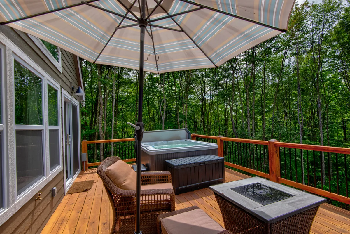 An inviting outdoor deck area featuring a bubbling hot tub, comfortable seating, and classic mountain railings. The view extends to the surrounding trees and mountains, highlighting a perfect spot to relax after a day of adventure.