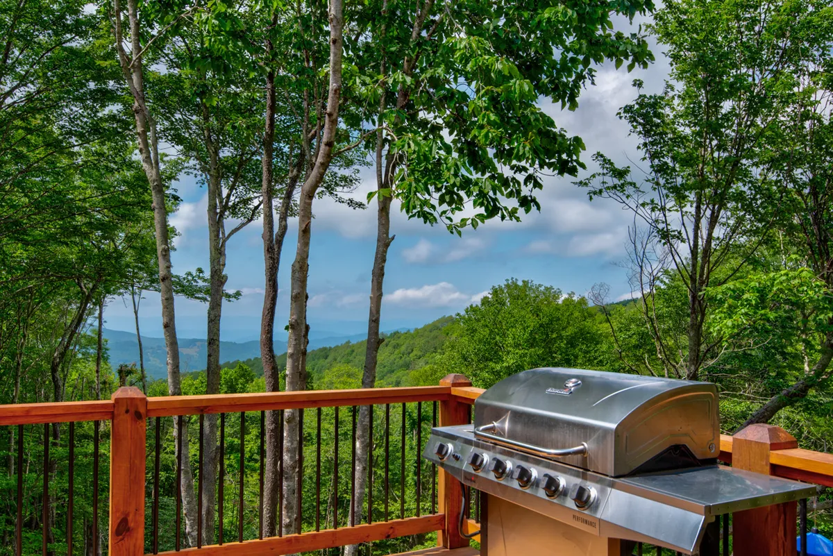 An exterior shot of the back deck with a grill and outdoor seating, surrounded by trees and with stunning mountain views. The scene suggests space for family gatherings, outdoor meals, and relaxation, with a refreshing mood that hints at both celebration and quiet moments.