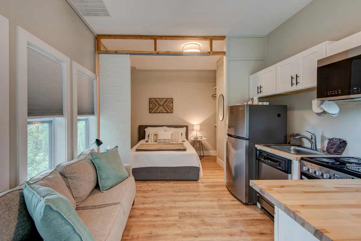 This image shows a comfortable mother-in-law suite with a bed, sofa, compact kitchen area, and decorative artwork. Natural light brightens the space, and the arrangement highlights how the suite combines sleeping, relaxing, and cooking areas in one inviting room.