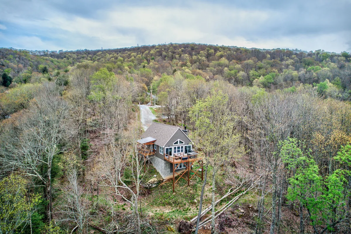 The image shows a beautiful, modern house nestled among tall trees atop Beech Mountain. The home's striking architecture stands out against the peaceful forest backdrop, with a spacious driveway leading up to the entry. The wraparound deck and large windows offer glimpses of panoramic mountain views, inviting guests to relax and take in sunsets or morning coffee surrounded by nature.