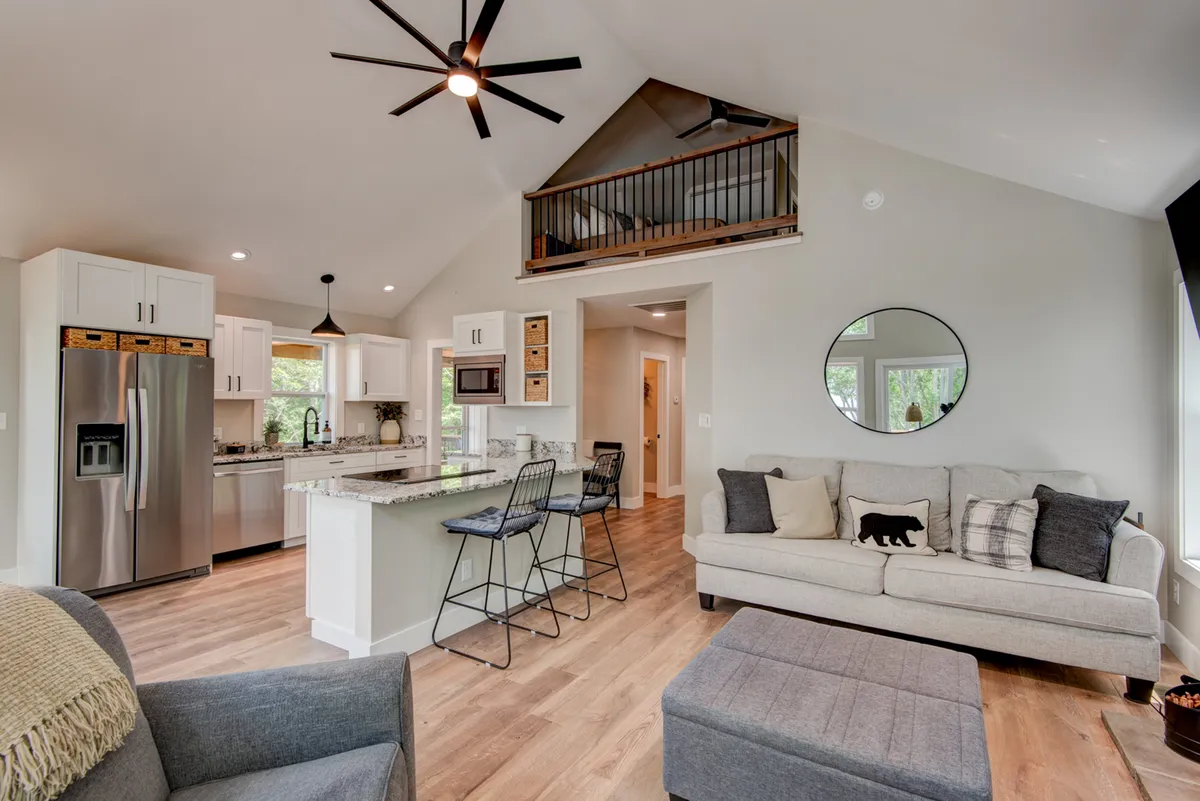 This interior shot captures the open-concept living area, showcasing the comfortable living room seating, high ceiling with a ceiling fan, open kitchen with modern appliances, and a dining area with barstools. Natural light pours in, emphasizing the spaciousness and connection between the great room, kitchen, and dining spaces.