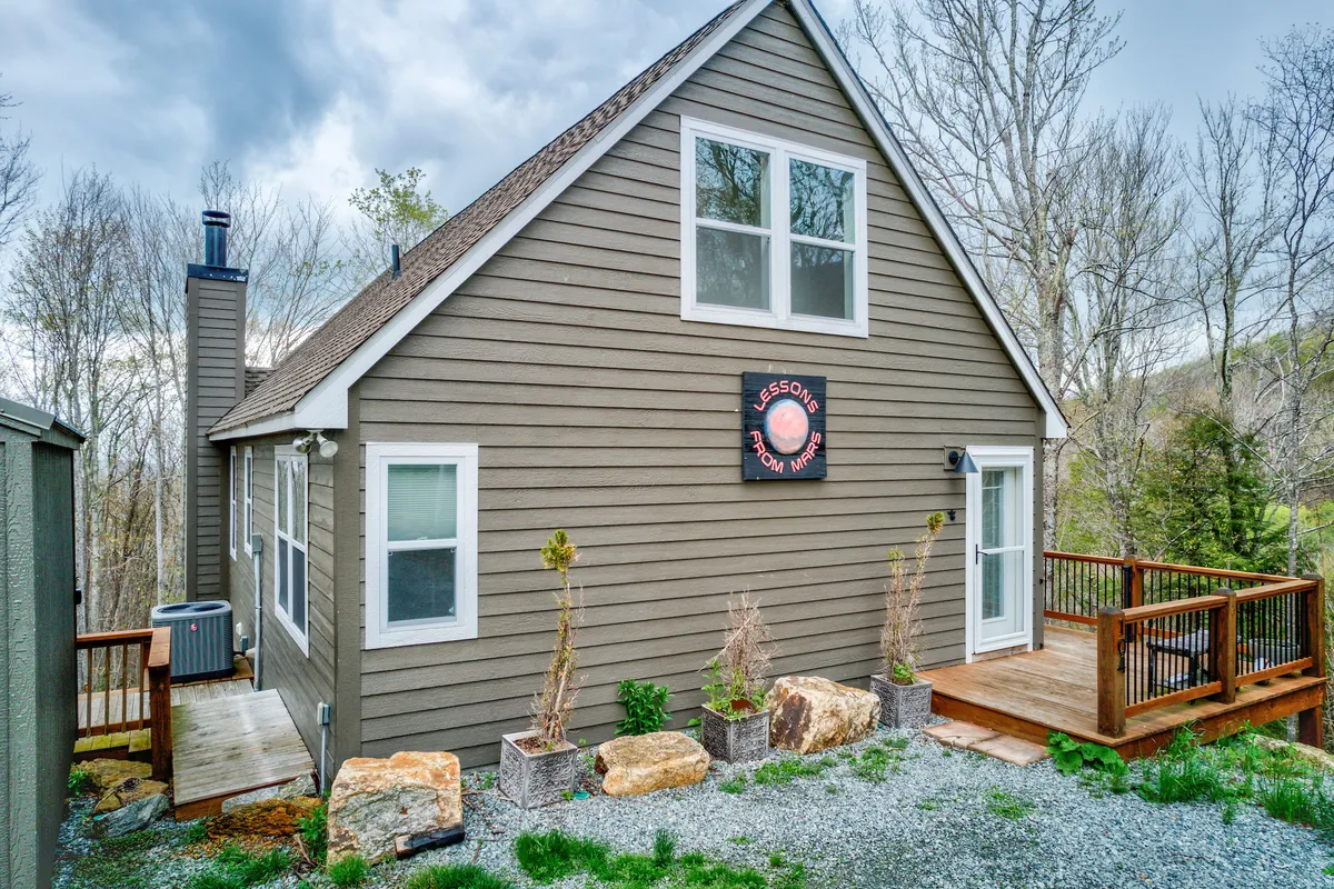 A wide exterior perspective of the house, showcasing unique architecture, a spacious deck, and a backdrop of lush trees. The image captures the whole home, emphasizing its setting and the opportunities for both togetherness and personal retreat within the property.