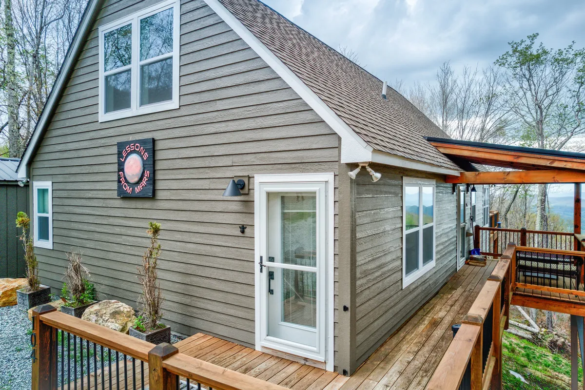 This image features the front entrance of the house, showcasing a spacious wooden deck with comfortable seating and a panoramic mountain view in the background. The sign near the door reads 'Lessons from Mins,' firmly identifying the property and establishing its unique context among the surrounding landscape.