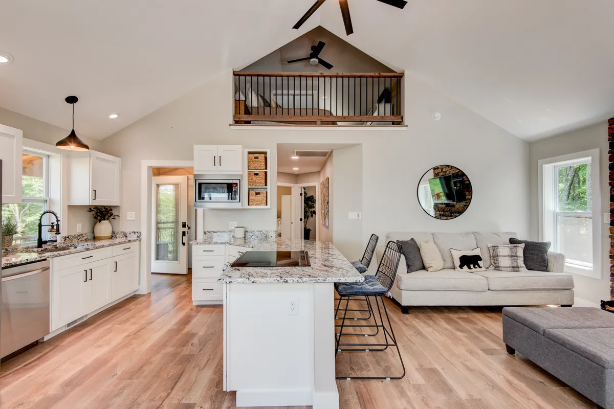 A contemporary kitchen with a granite island is connected to a comfortable sofa and an open loft space above. The kitchen is well-equipped with stainless appliances and the layout encourages casual interaction across spaces.
