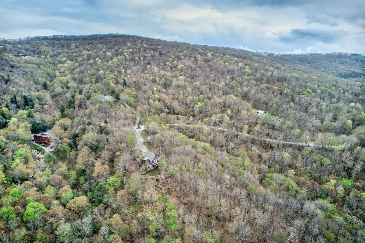 A serene, wide-angle view of scenic mountains with an isolated cabin and a winding road in the foreground. The scene showcases the vast, natural landscape surrounding the home, with hints of autumn foliage and the promise of hiking, exploration, and stargazing.