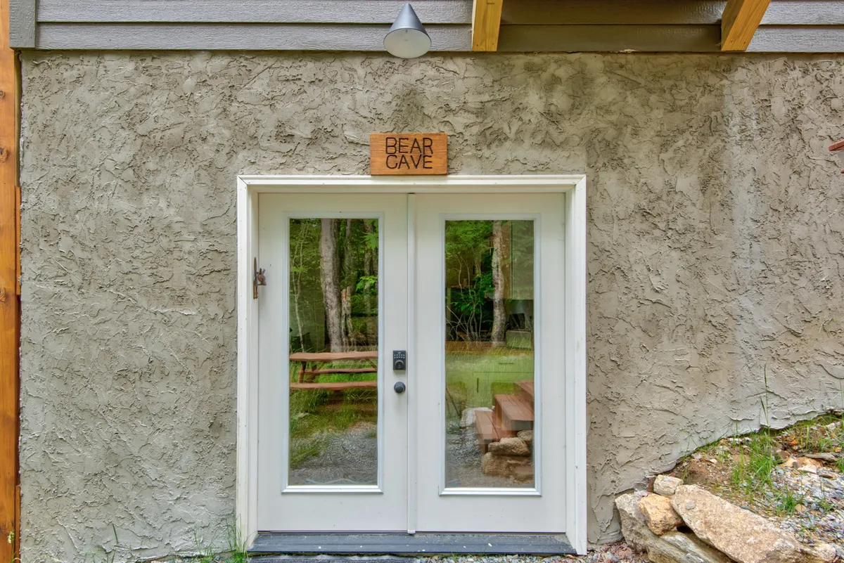 A cozy exterior with double doors, a 'BEAR CAVE' sign, and a natural setting that hints at both adventure and a welcoming retreat. The surrounding greenery and inviting entrance suggest a perfect base camp for exploring nearby attractions.