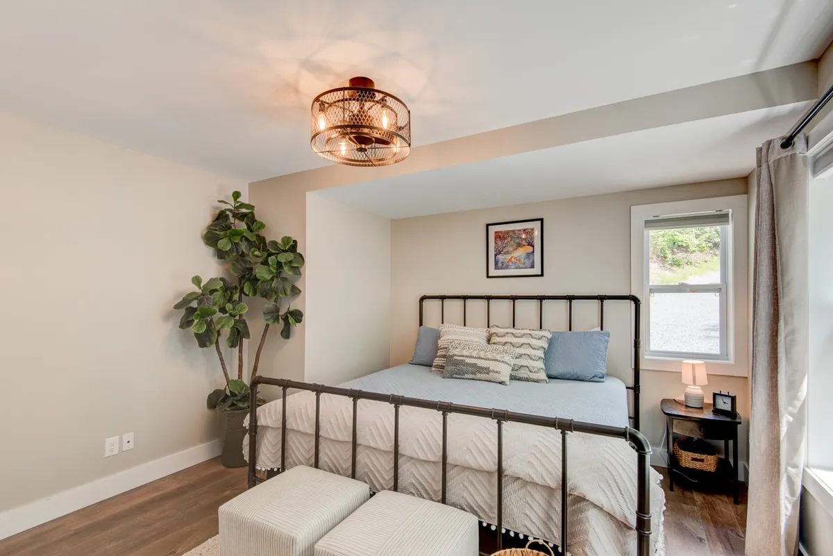 A tranquil bedroom scene with a neatly made bed, nightstand, potted plant, a window allowing morning light to filter in, and soft lighting—a peaceful invitation to rest and rejuvenate.