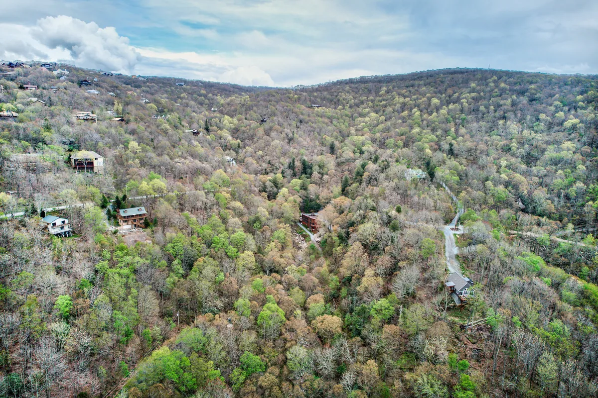 A wide, tranquil view of lush green trees, distant hills, scattered houses, and a winding road weaving through the landscape. The image evokes the expanse and quiet beauty of the surrounding mountains, highlighting the sense of adventure and nature just outside the doorstep.