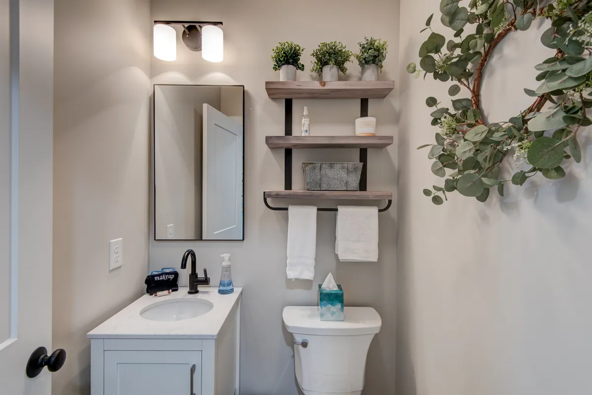 A serene bathroom setting with a vanity sink, mirror, open shelving featuring decor and towels, and touches of greenery. The space appears private and inviting, ideal for personal refreshment.