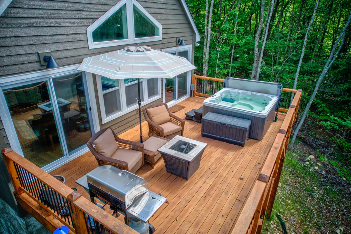 This inviting exterior shot showcases the hot tub, an outdoor seating area, grill, and umbrella, all arranged on a spacious deck. The setting is perfect for cozying up, sharing a meal, or enjoying starlit evenings, capturing both the social and romantic possibilities this retreat offers.